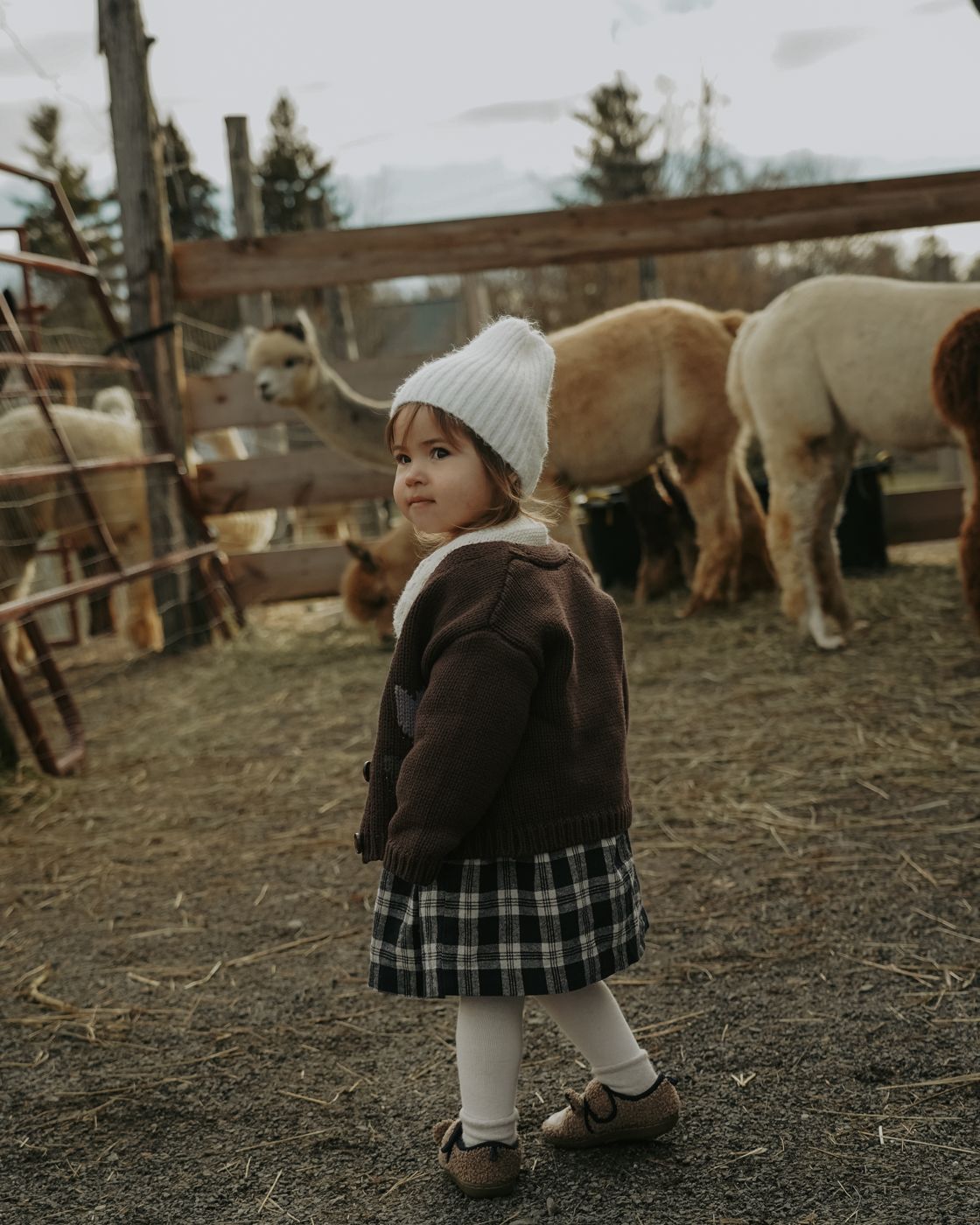 Toddler in a brown cardigan looking back over her shoulder at the farm