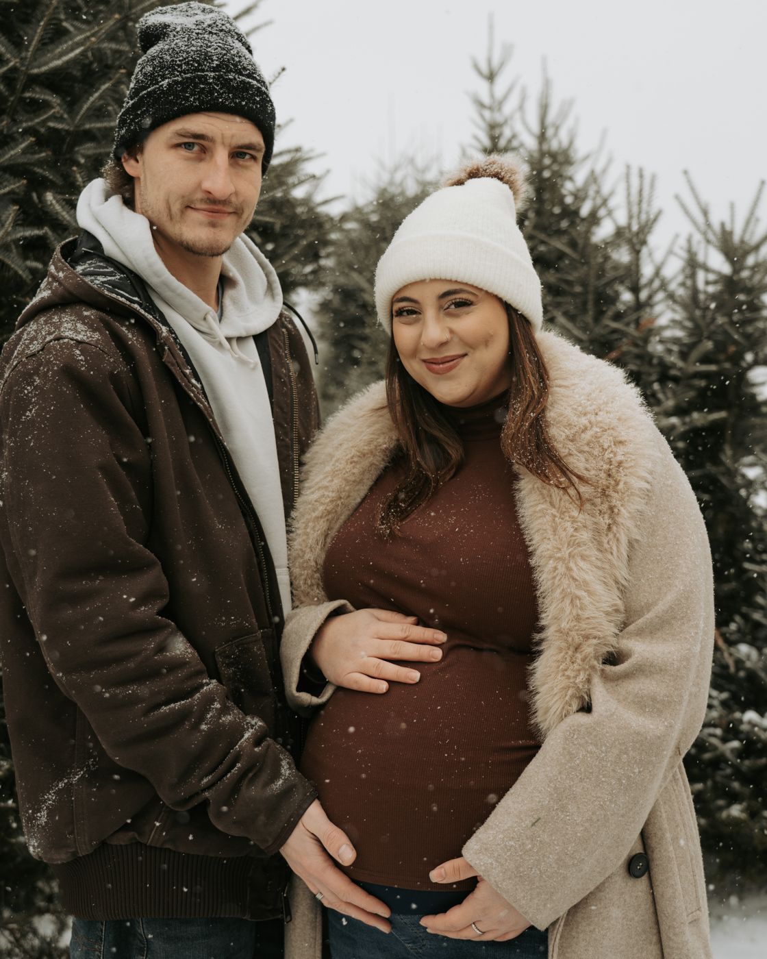Expecting couple in winter coats smiling among snowy evergreens