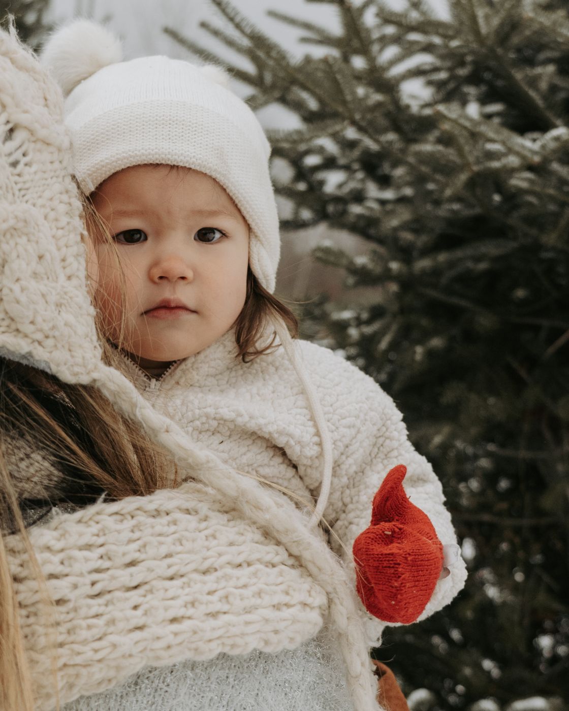 Toddler in a white knit hat and red mittens held close by mom