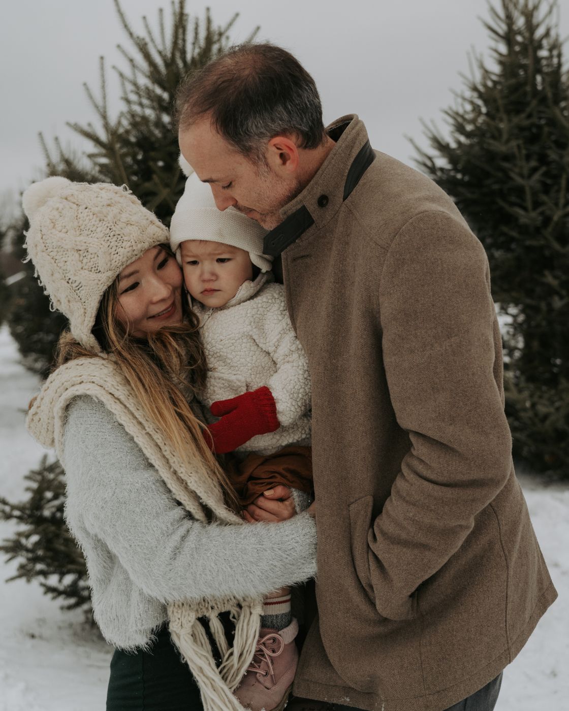 Parents hugging their toddler close among snowy Christmas trees