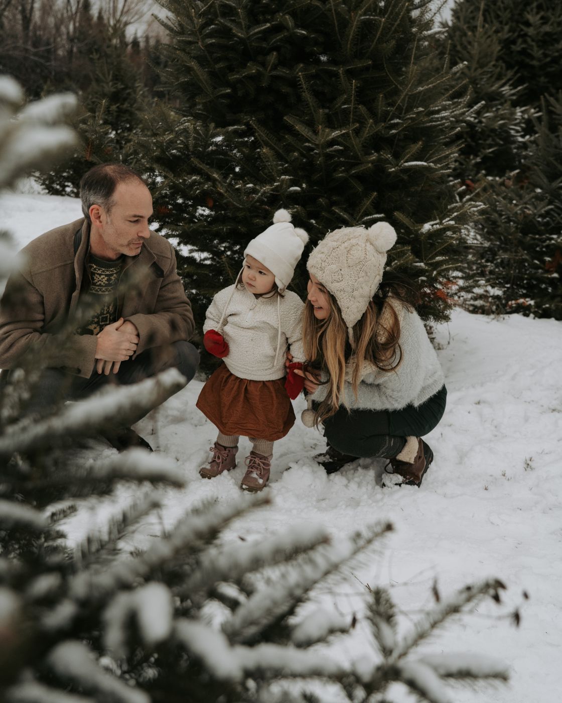 Parents crouching in the snow with their toddler between Christmas trees